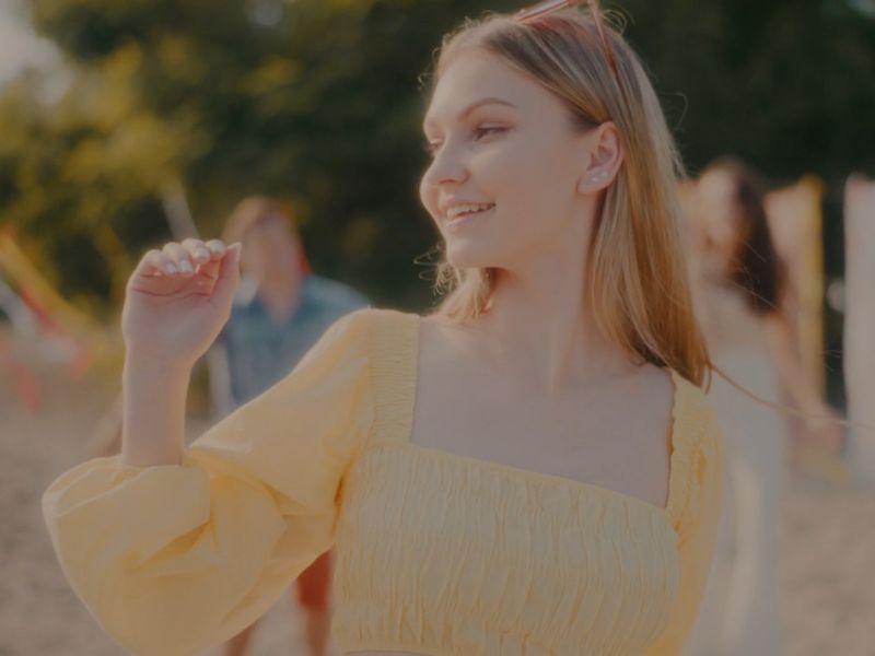  Mlada žena sa svetlom kosom u žutoj bluzi smeši se na otvorenom, okružena ljudima tokom letnje zabave.